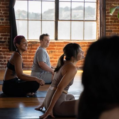 People seated on yoga mats in a sunlit brick studio near large windows.
