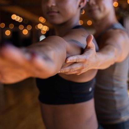 Two people in workout clothes balancing in a partner yoga pose with arms extended, warm lights in the background