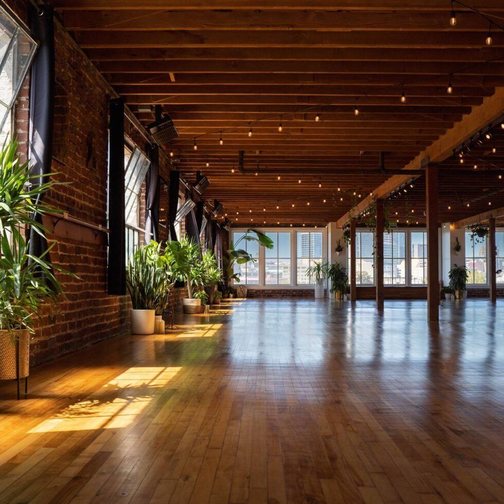 Sunlit empty loft with wooden floors, exposed beams, brick walls, and potted plants