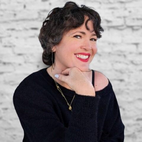 Smiling person in a black sweater, resting chin on hand against a white brick wall.