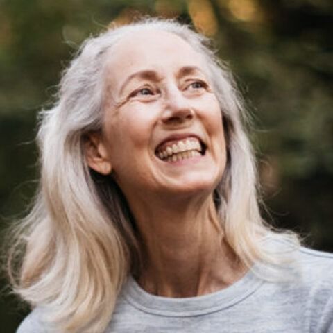 Smiling woman with long gray hair looking upward outdoors in a gray shirt