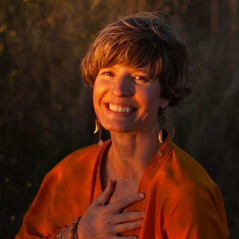 Smiling person in an orange robe at sunset, hand on chest, outdoors