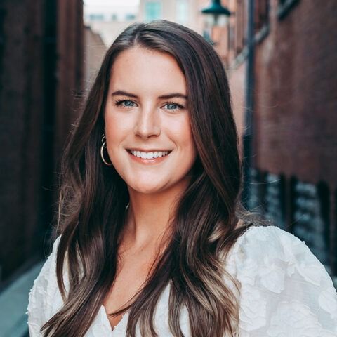 Smiling person with long brown hair in a white blouse standing on a city street
