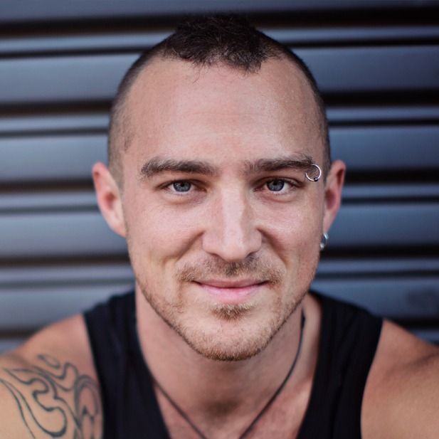 Close-up portrait of a smiling person with a shaved head and eyebrow piercing against a gray striped background