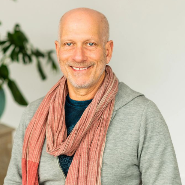 Smiling bald man in a gray cardigan and pink scarf indoors with a plant in the background