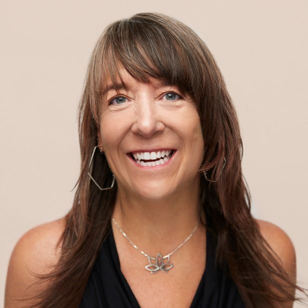 Smiling woman with long brown hair against a beige background, wearing a black top and silver necklace