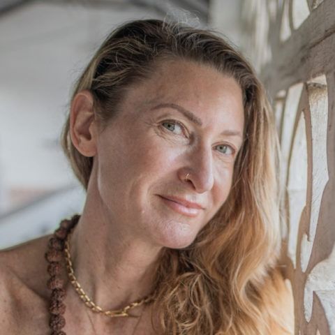 Close-up portrait of a woman with long hair leaning by a sunlit window, wearing a gold necklace.