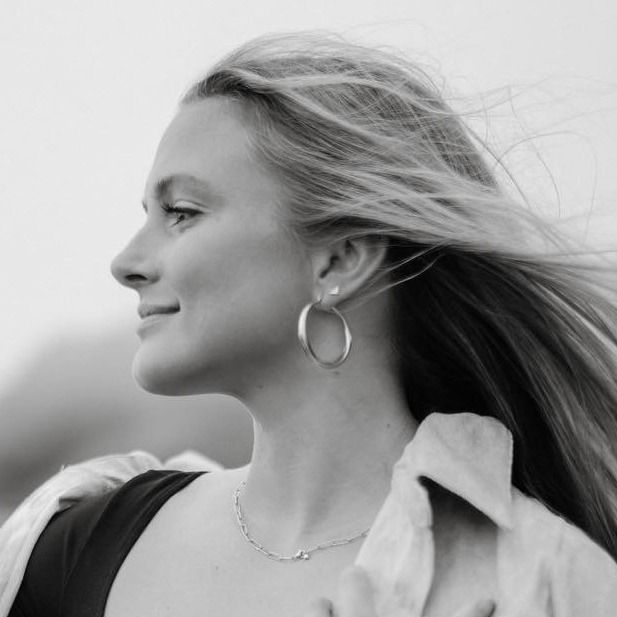 Woman in profile with windblown hair, wearing hoop earrings and a collared shirt, against a soft sky backdrop