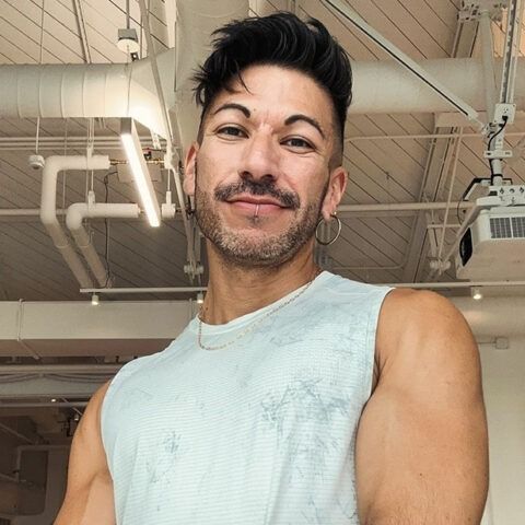 Smiling person in a sleeveless light blue top, posing in a bright gym interior.