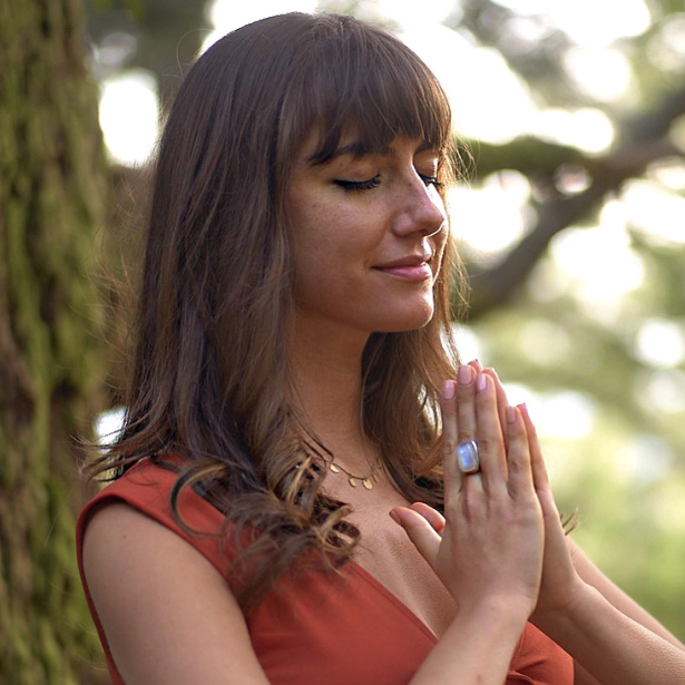 Woman in a rust-colored top with hands in prayer, eyes closed outdoors near a tree.
