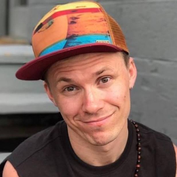 Smiling person in a colorful cap and black shirt, tilting their head against a gray background