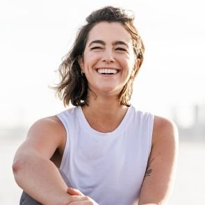 Smiling woman in a white sleeveless top outdoors, with arms crossed against a bright sky.