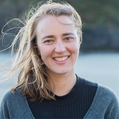 Smiling person with windblown hair in a dark sweater outdoors by water