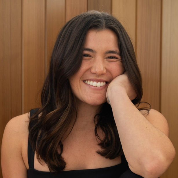 Smiling woman with dark hair resting her head on her hand against a wooden backdrop