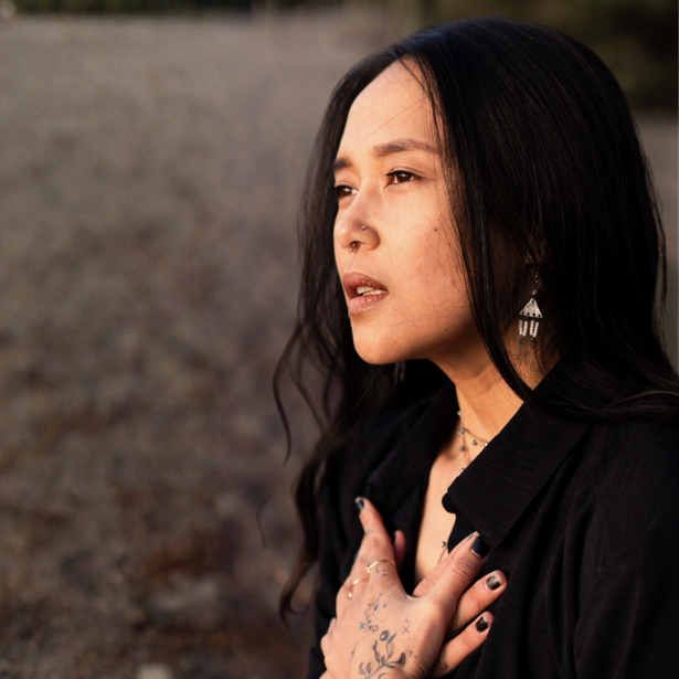 Woman in a black shirt, shown in profile with hands on chest, standing outdoors at sunset.