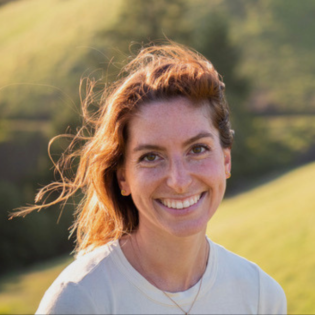 Smiling woman outdoors with windblown hair in warm sunlight, with green hills behind her
