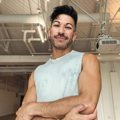 Person doing a yoga boat pose on a mat in a bright studio, smiling toward the camera.