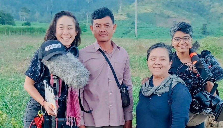 Four people smiling, outdoors, camera and audio equipment, green landscape background.