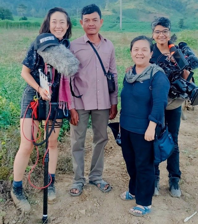 Four people in a field, smiling. Two hold cameras, one has a microphone, and one appears to be the subject.