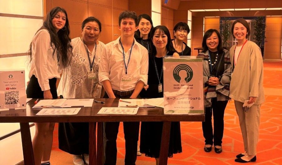 Group of people at a registration table; smiling, mixed genders and ethnicities; indoor setting with orange carpet.