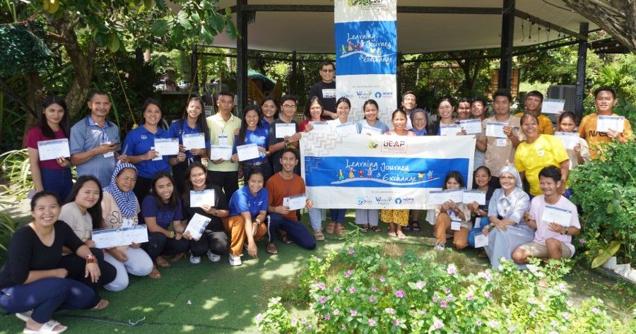 A group of people standing outdoors under a banner for a graduation or training ceremony, holding certificates.