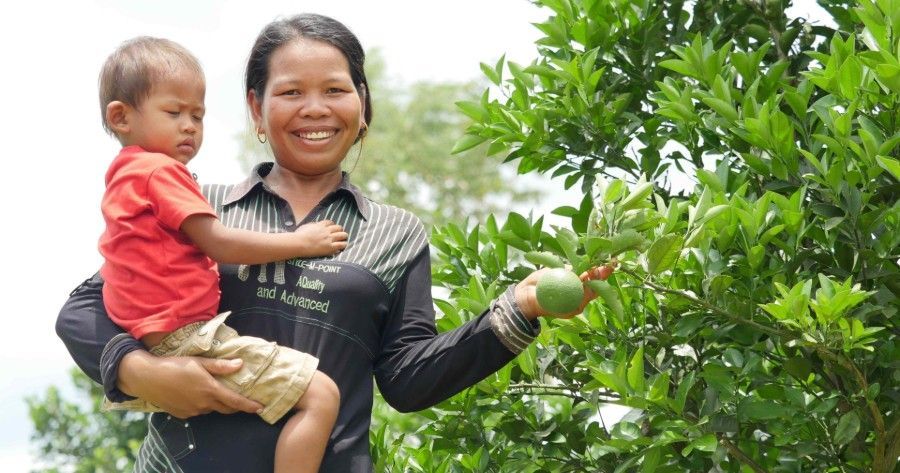 Smiling person holding a child beside a fruit tree, reaching toward hanging green fruit.