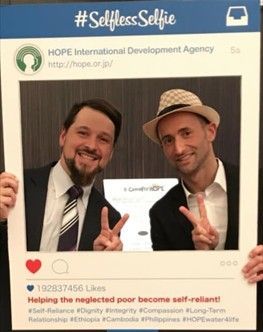 Two men holding a selfie frame with an organization's logo. Text says: #Selfless Selfie, and