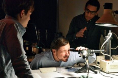 Three people leaning over equipment in a dimly lit lab; one person peers through a device, others observe.