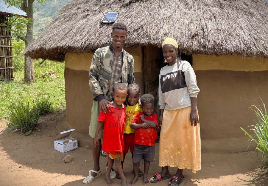 Family standing outside a thatched hut, with two children in red shirts and two adults posing together.