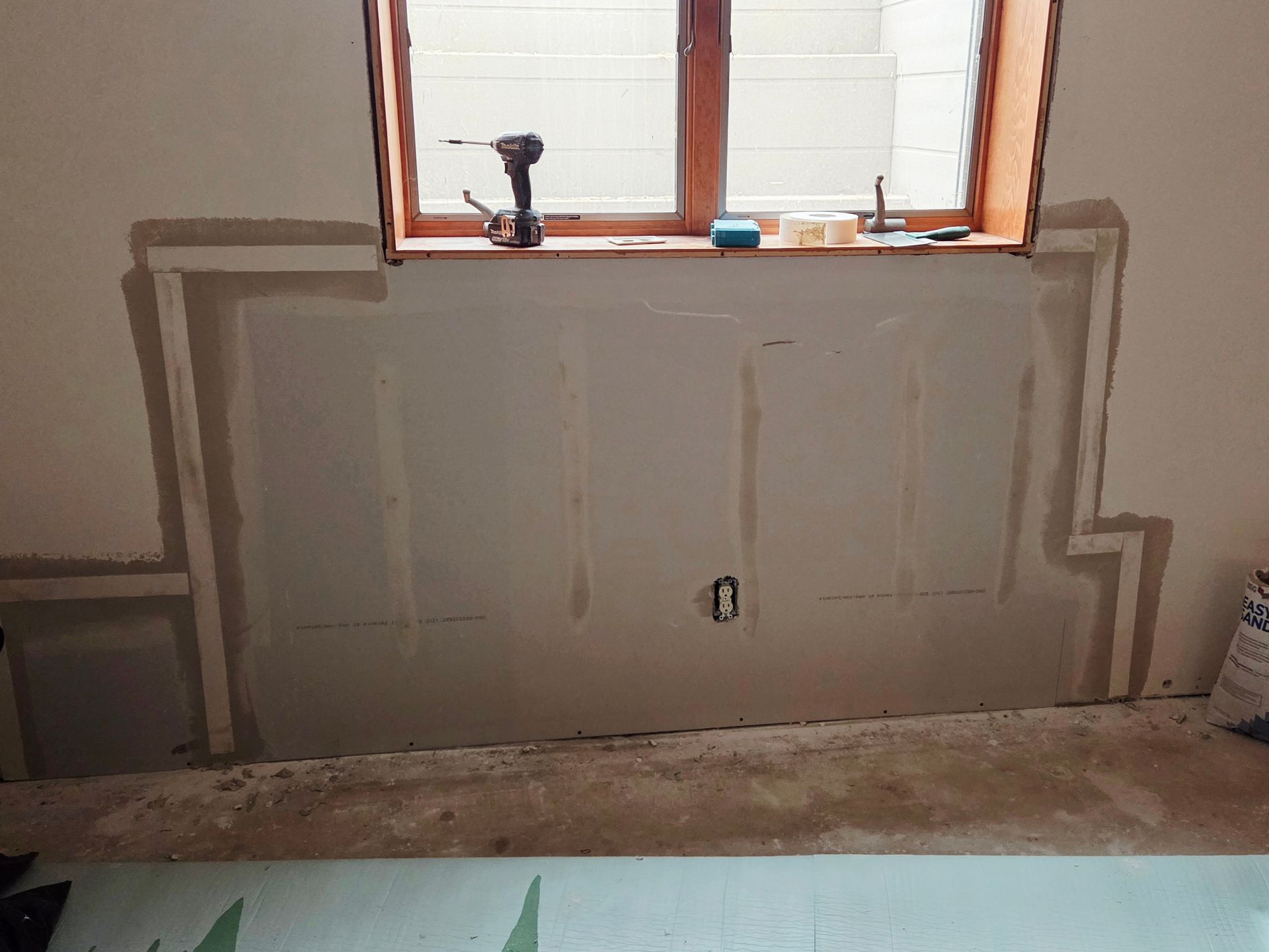 Drywall patches and joint compound applied to a wall beneath a window, with a drill sitting on the window sill.