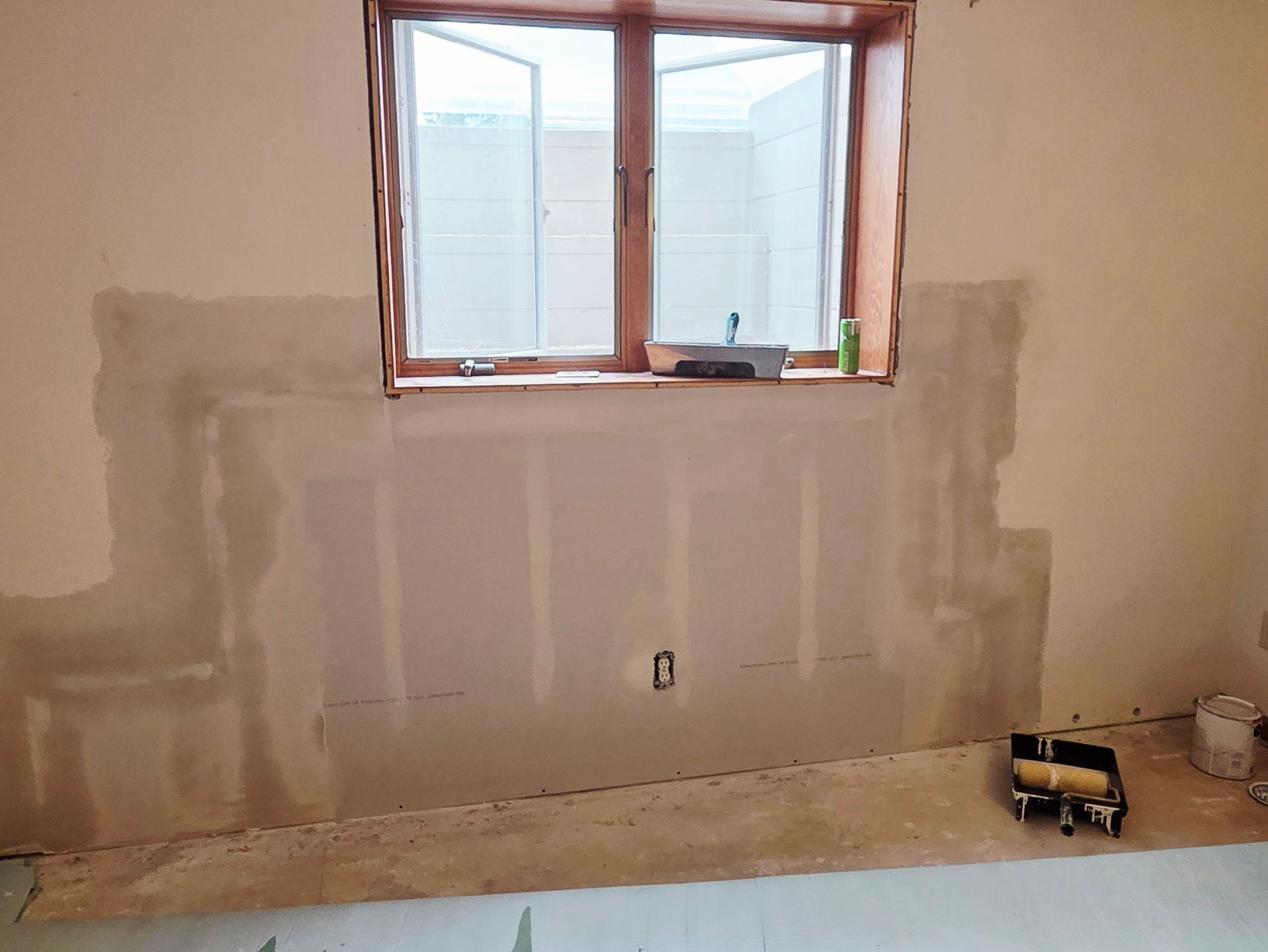 Interior wall section with newly installed drywall and taped seams below a wooden window frame in a room under renovation.