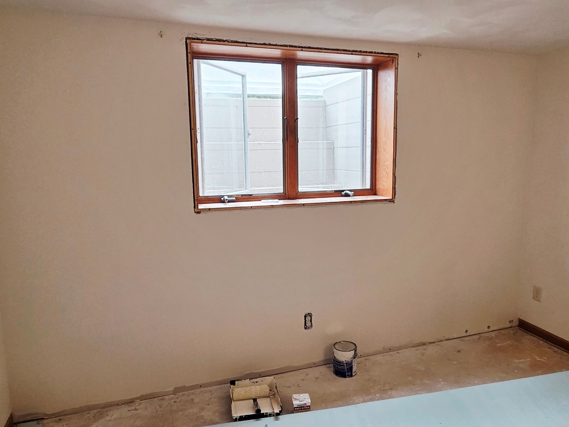 An interior room with beige walls, a central window with a wooden frame, an electrical outlet, and debris on the floor.