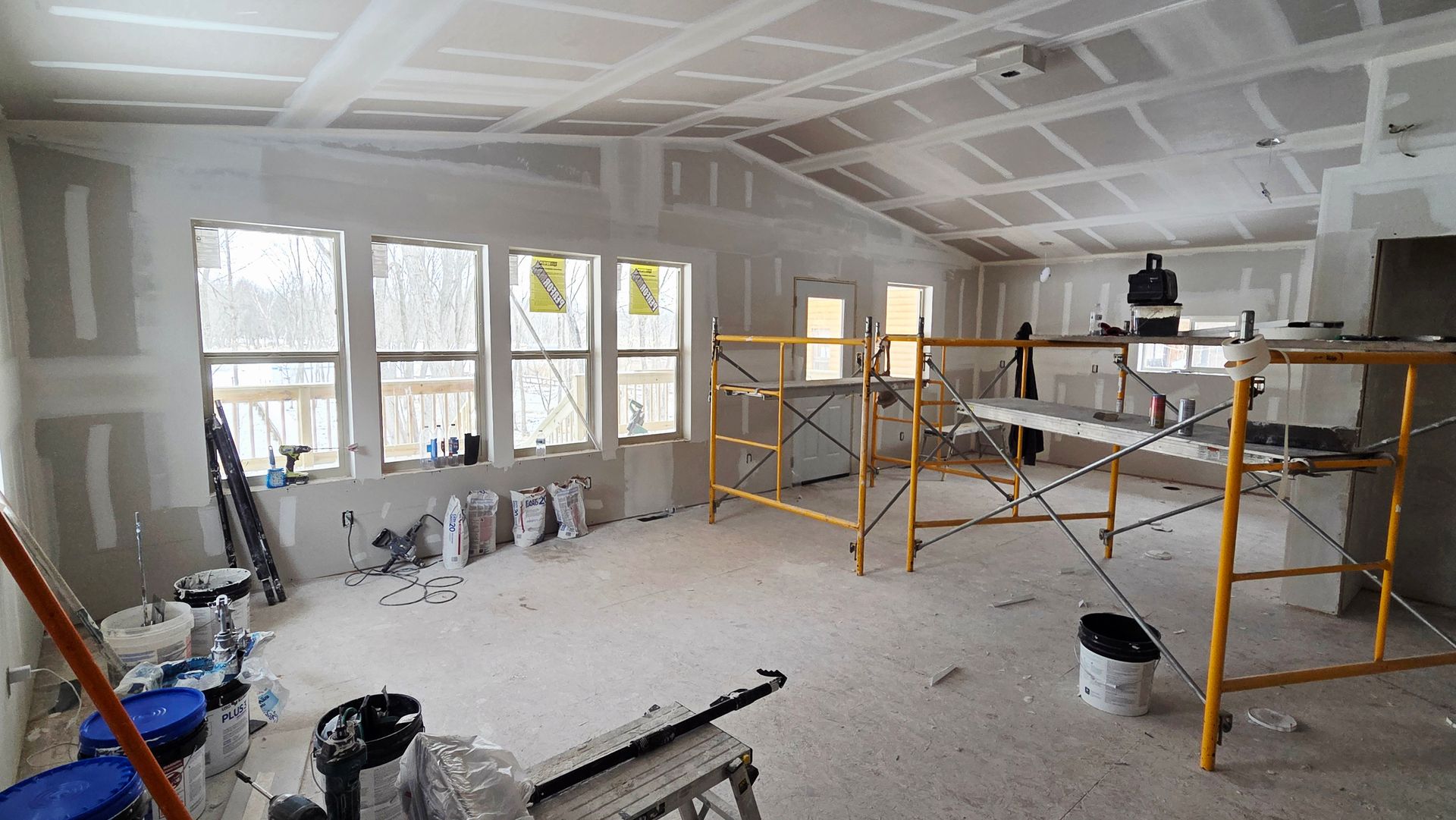 Interior view of a room under construction with white drywall, windows, and yellow metal scaffolding on a dusty floor.