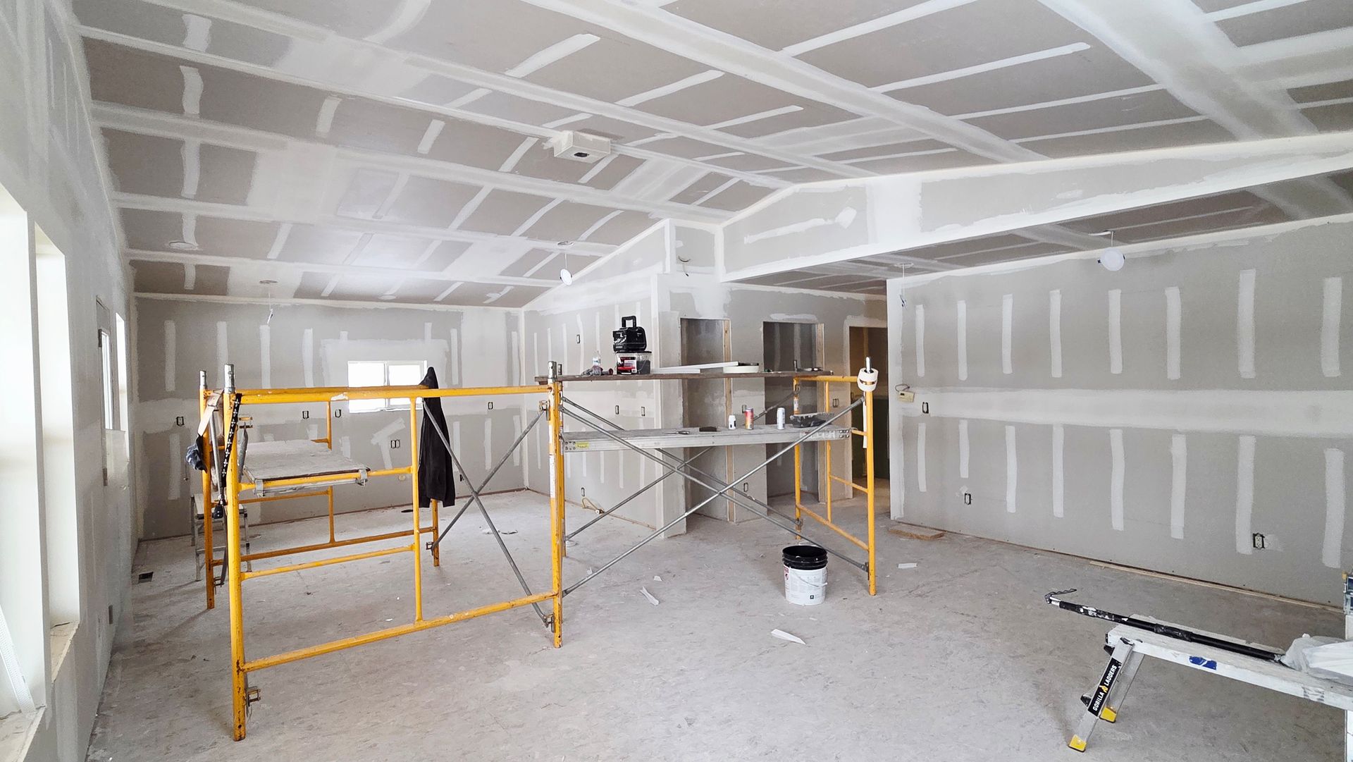 An interior room under construction with drywall installed on walls and vaulted ceilings, featuring scaffolding.
