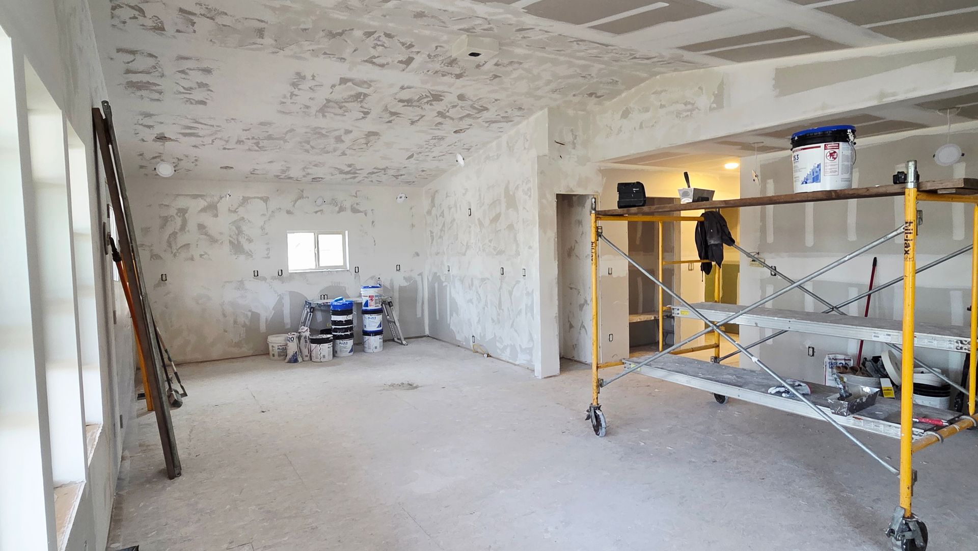 An interior room under construction with white drywall, a yellow scaffolding tower, and building supplies.