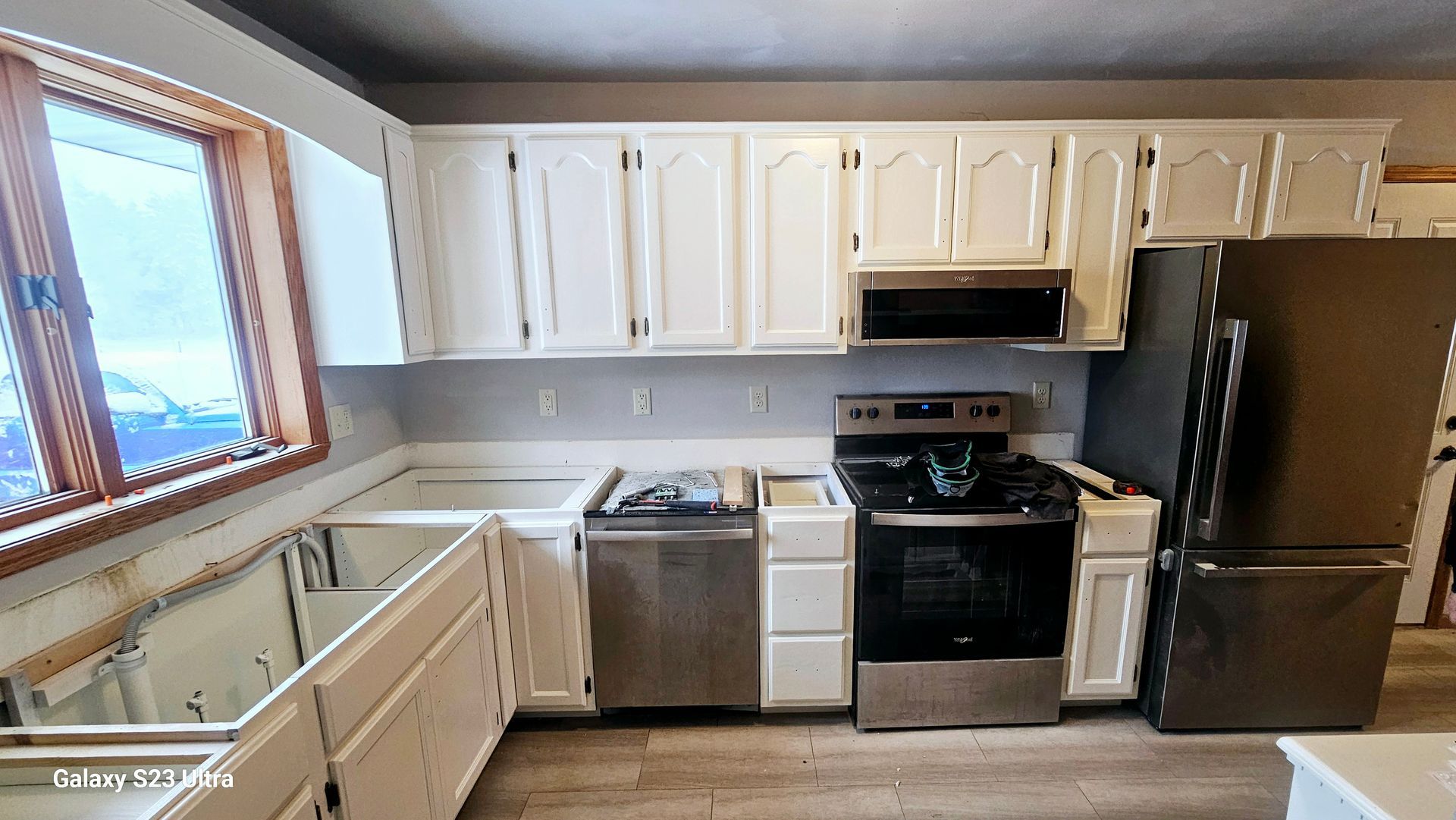 A kitchen with white cabinets, stainless steel appliances, a window on the left, and light wood-look flooring.
