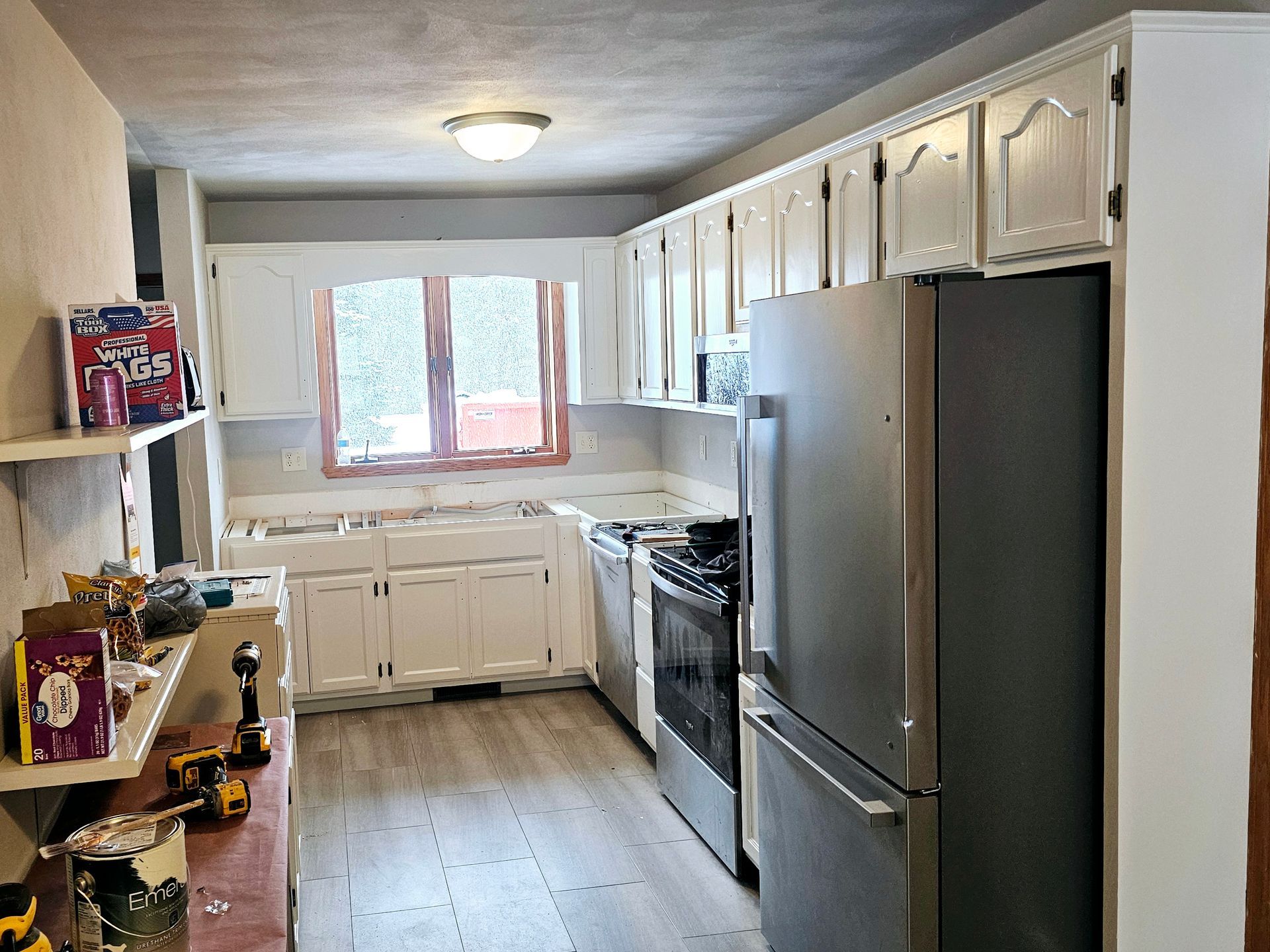 A kitchen under renovation with white cabinets, a stainless steel refrigerator, black stove, and light wood-look flooring.