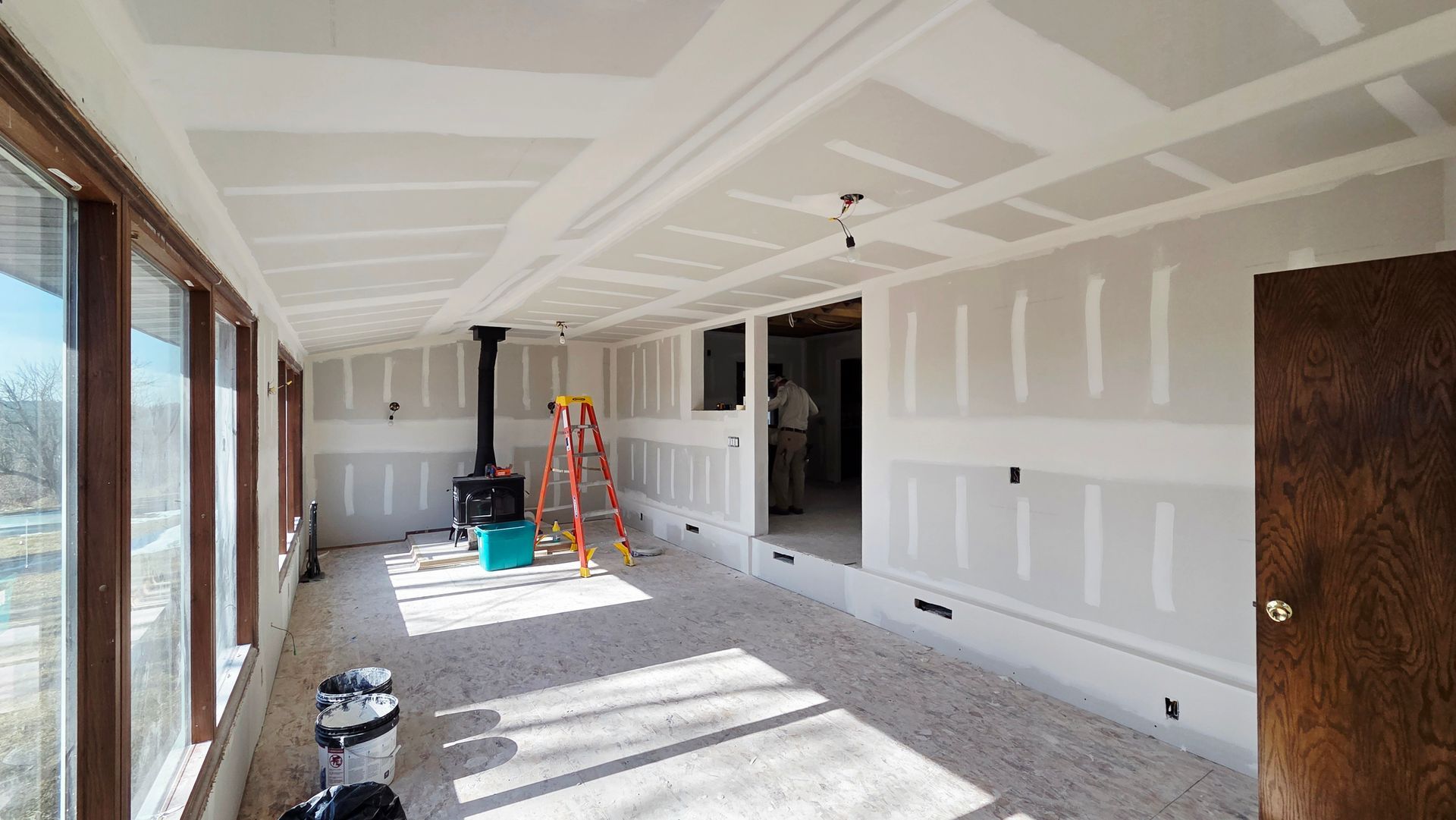 A room under construction with white drywall, a tall ladder, a wood-burning stove, and windows looking out onto a landscape.