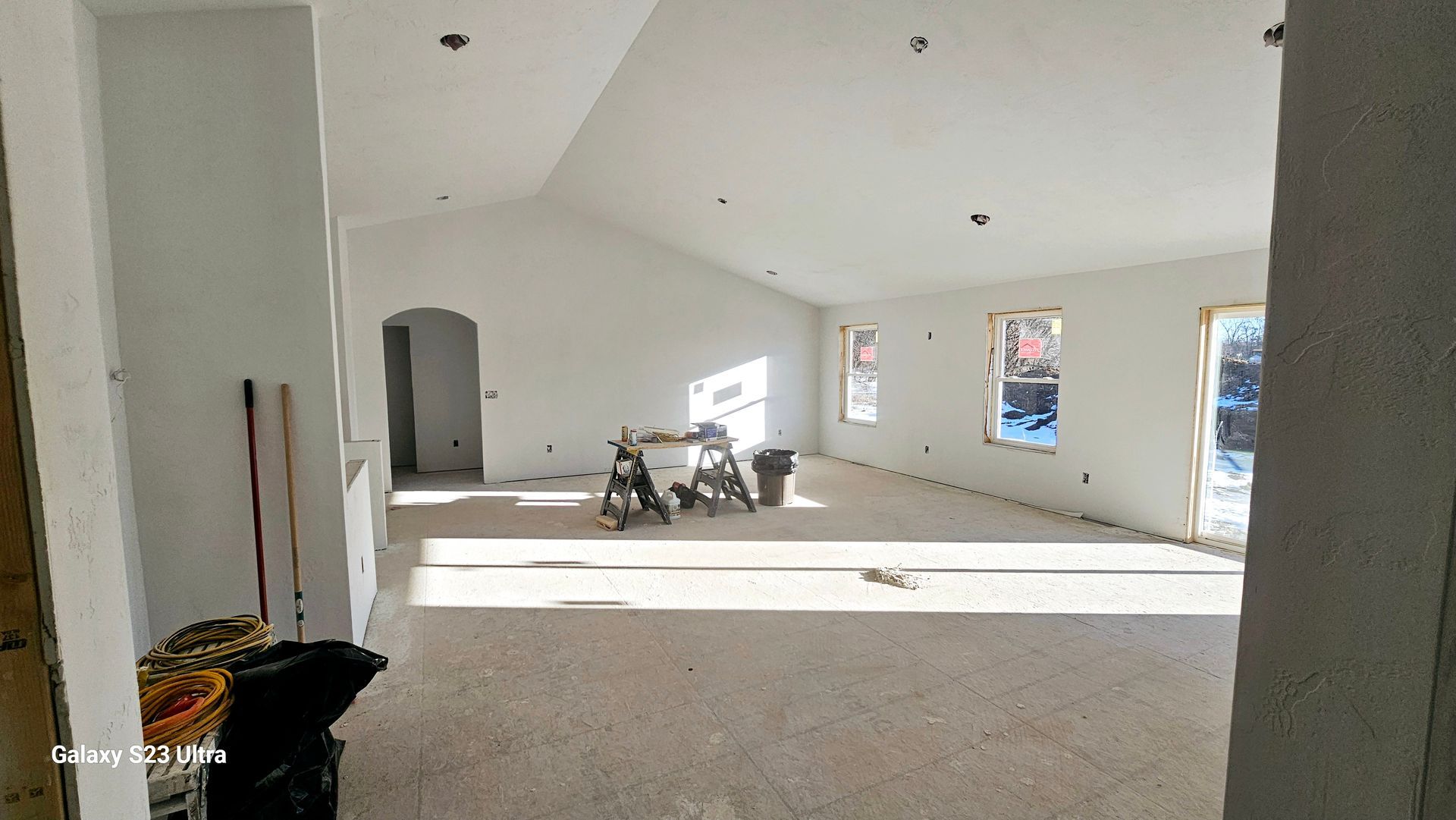 A bright, unfinished room with white walls, a vaulted ceiling, and three windows, currently containing construction gear.