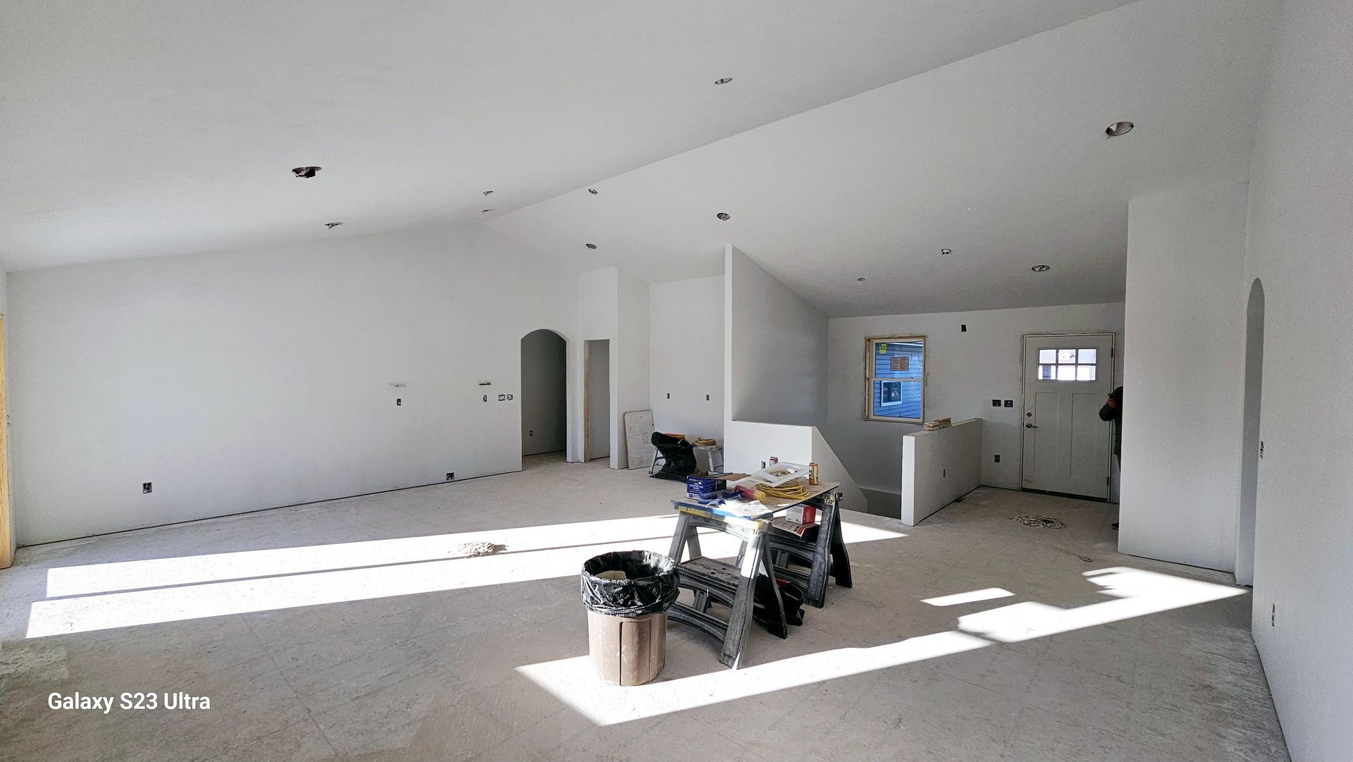 An interior view of a wide, unfinished white-walled room with vaulted ceilings, recessed lighting, and a construction bin.