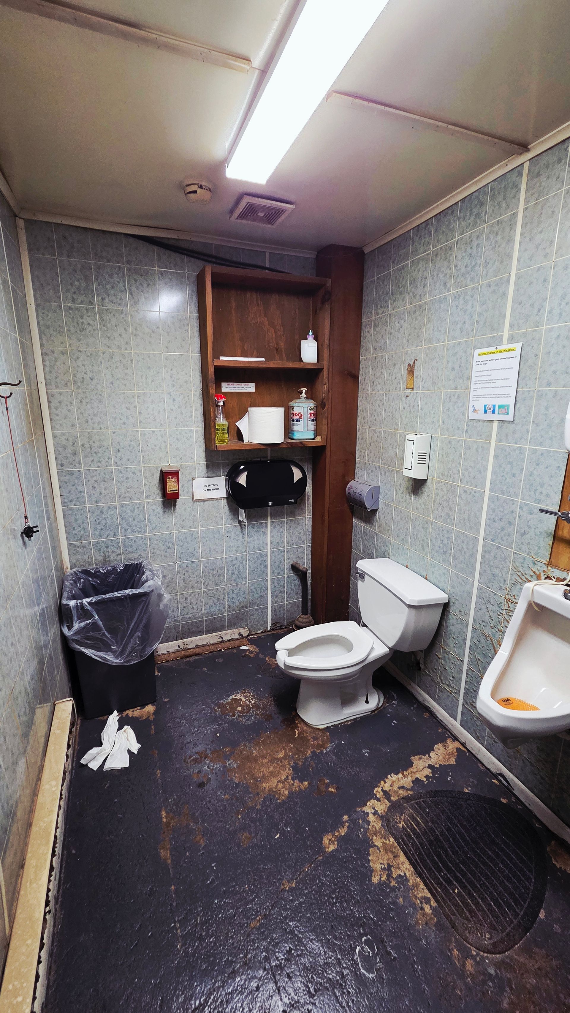 A small, dimly lit public restroom with a toilet, urinal, wooden shelving, and a dark, stained floor with scattered debris.