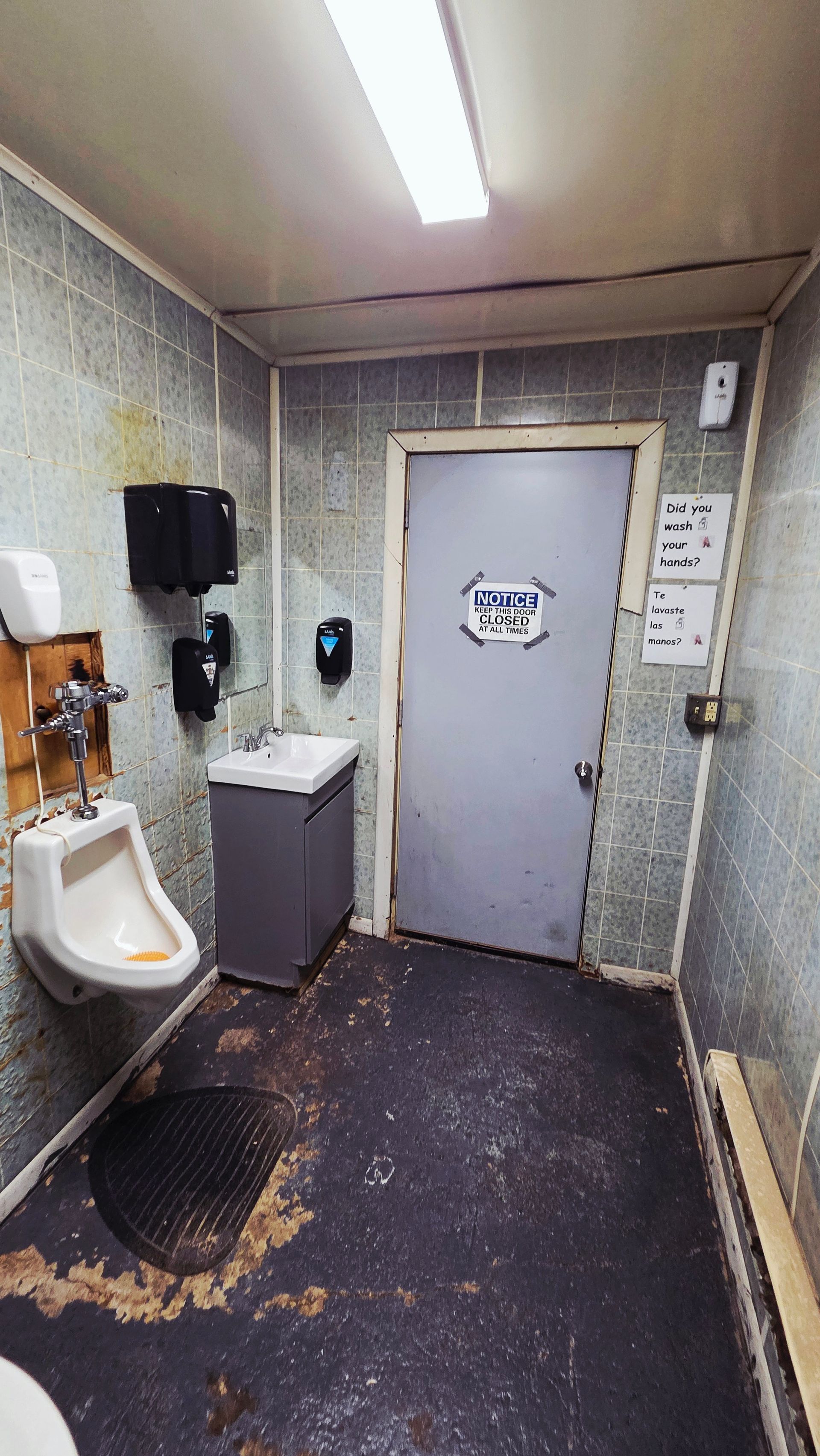 A small, worn public restroom featuring a white urinal, a grey vanity sink, and a door with a sign, all on a dark floor.