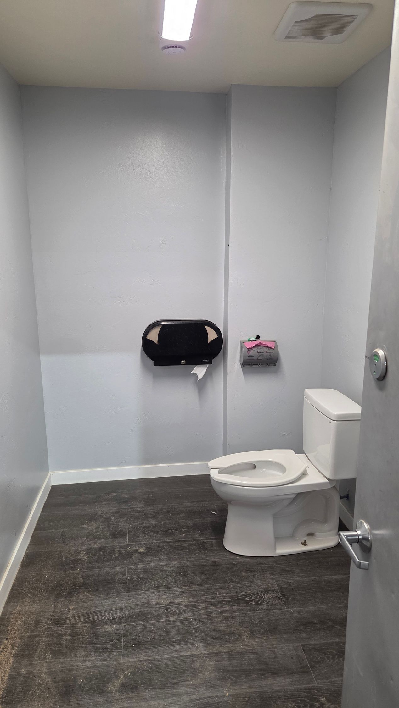 A white toilet in a small, grey-walled restroom with dark wood-look flooring and a wall-mounted toilet paper dispenser.