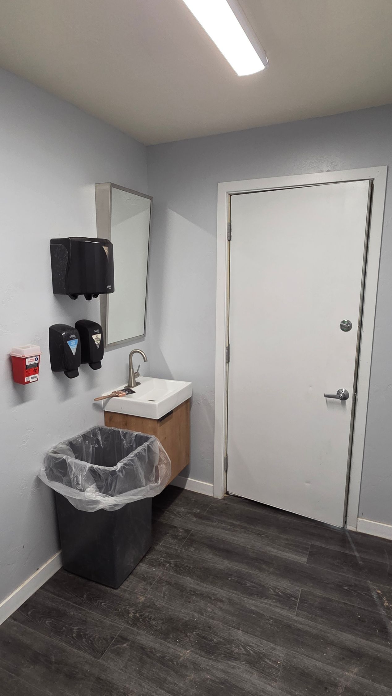 A restroom interior featuring a wall-mounted sink, mirror, trash can, soap dispensers, and a white door on a gray floor.