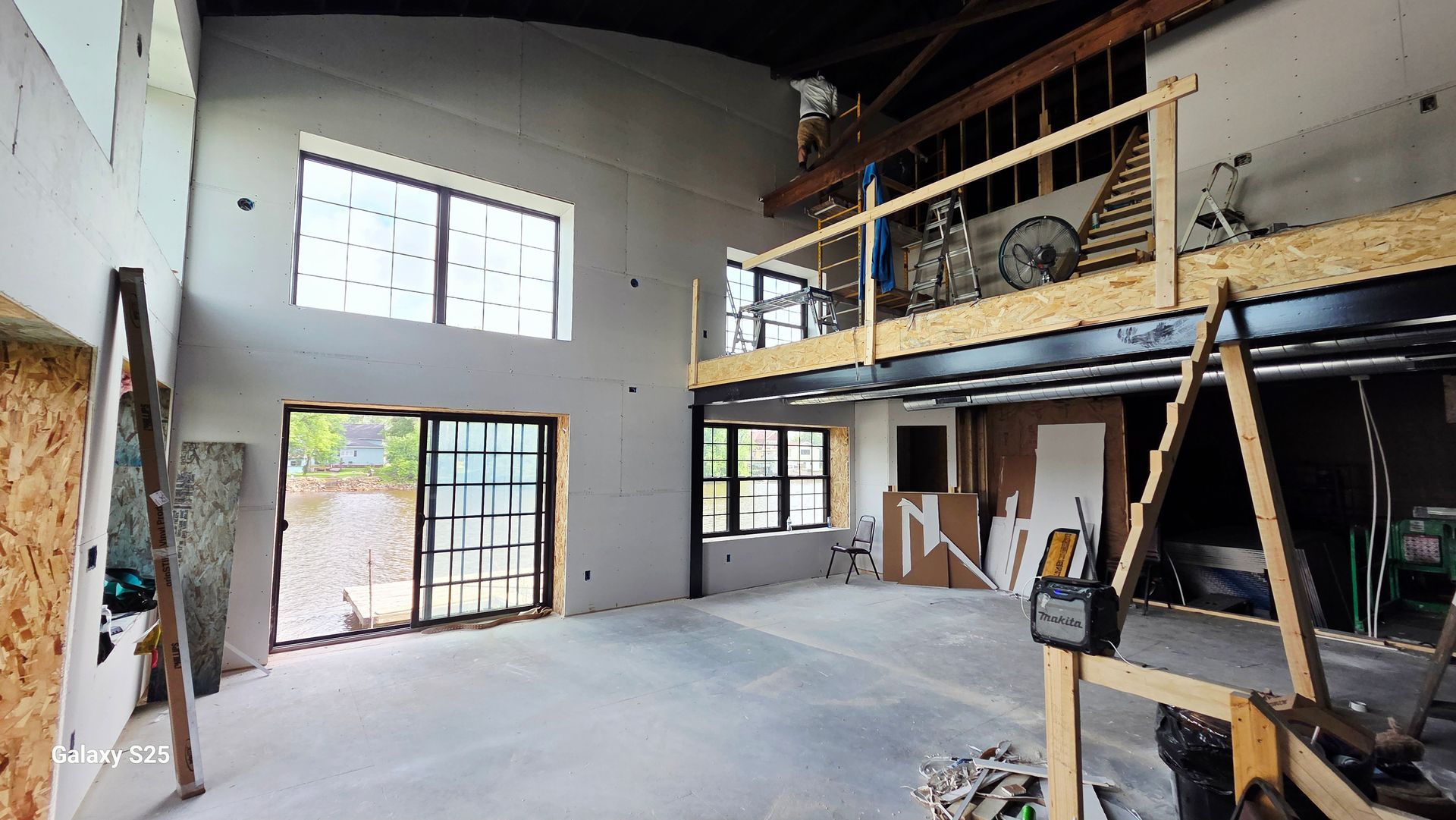 An interior view of a room under construction with a mezzanine level, large windows, and a ladder leaning against a wall.
