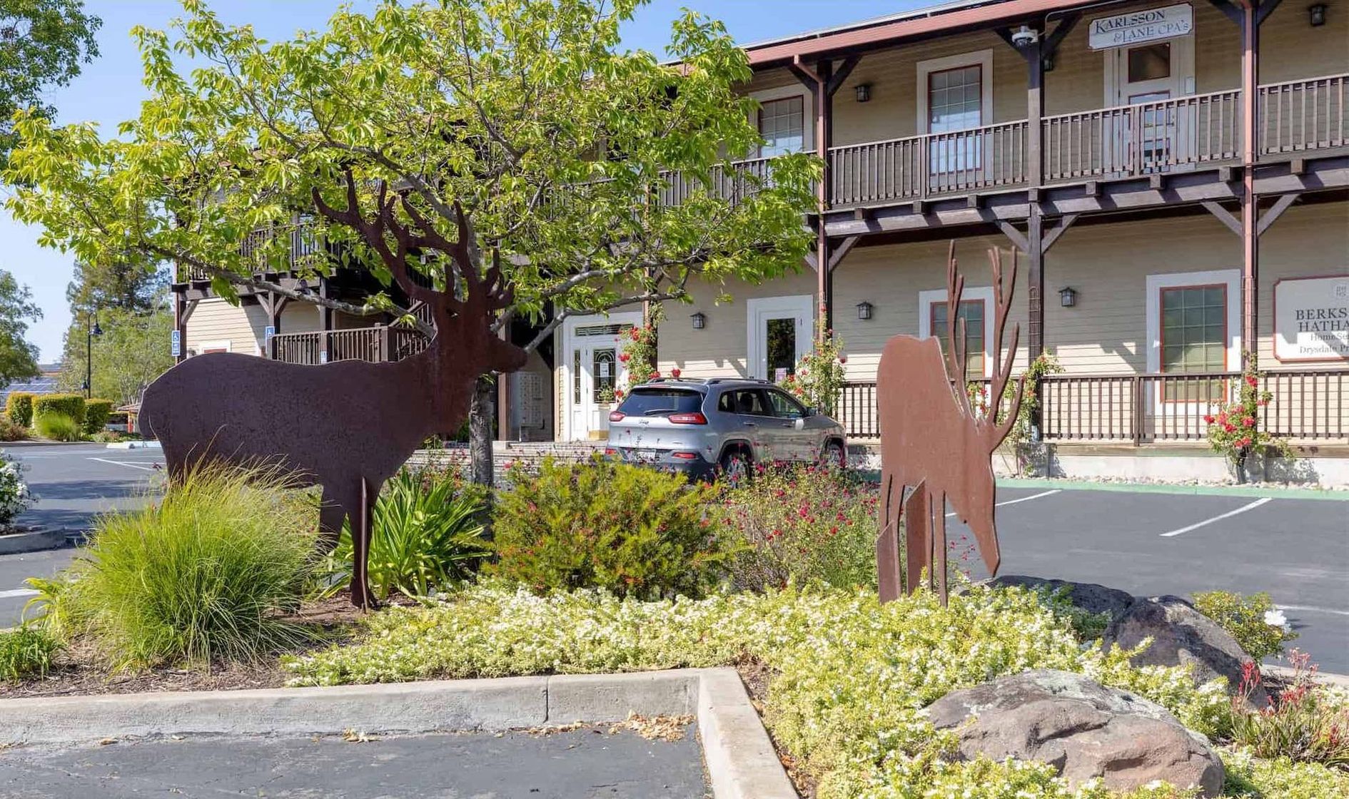 Metal deer sculptures stand in a landscaped garden in front of a two-story building with a parking lot.