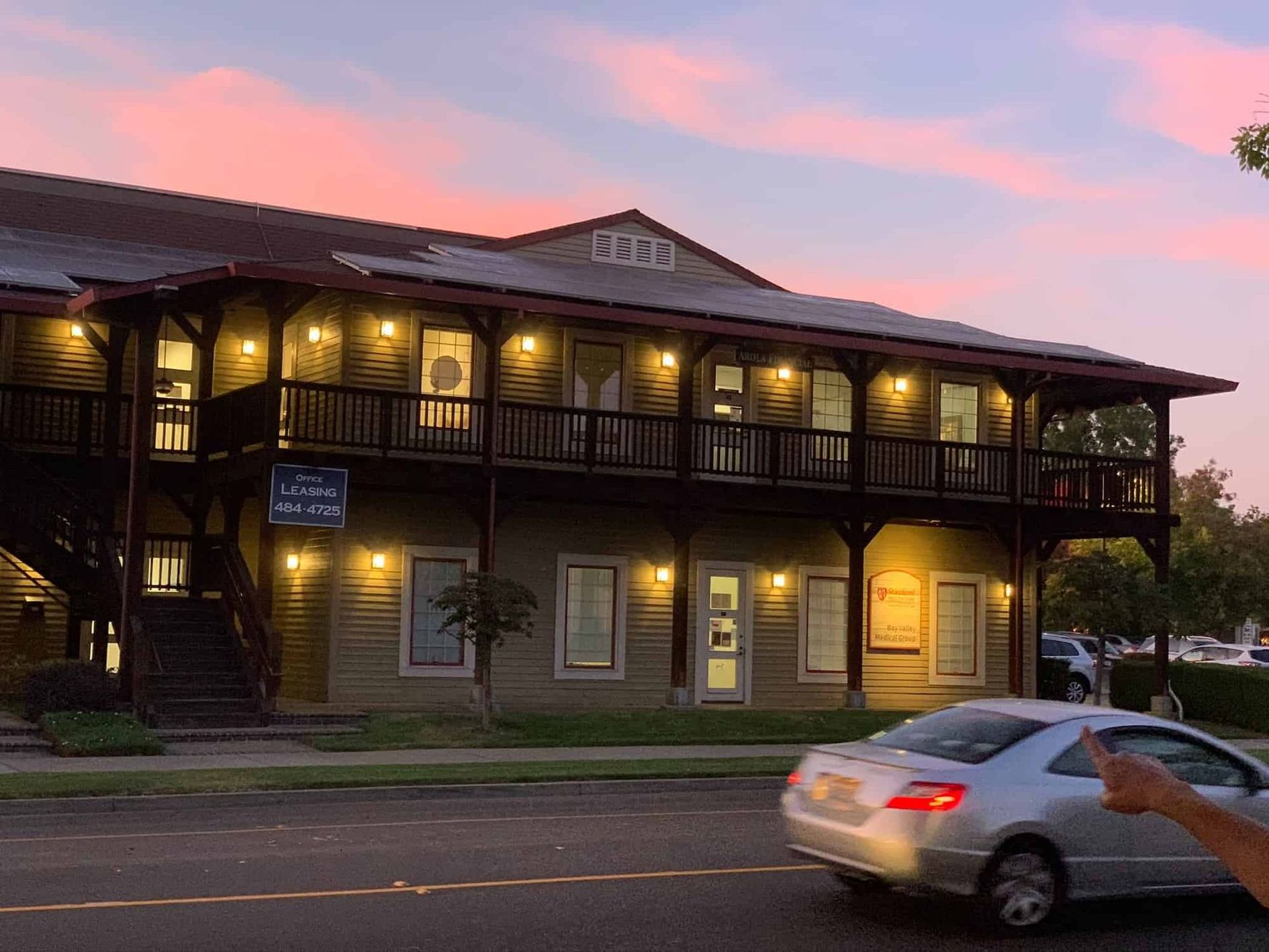A two-story building with a wooden balcony at dusk, with a silver car passing in the foreground.