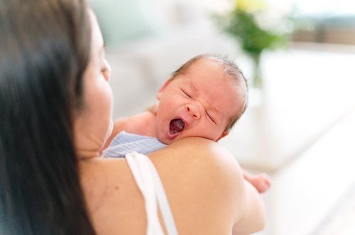 Baby yawning while held on a shoulder. Soft blue towel, white strap. Bright natural light.