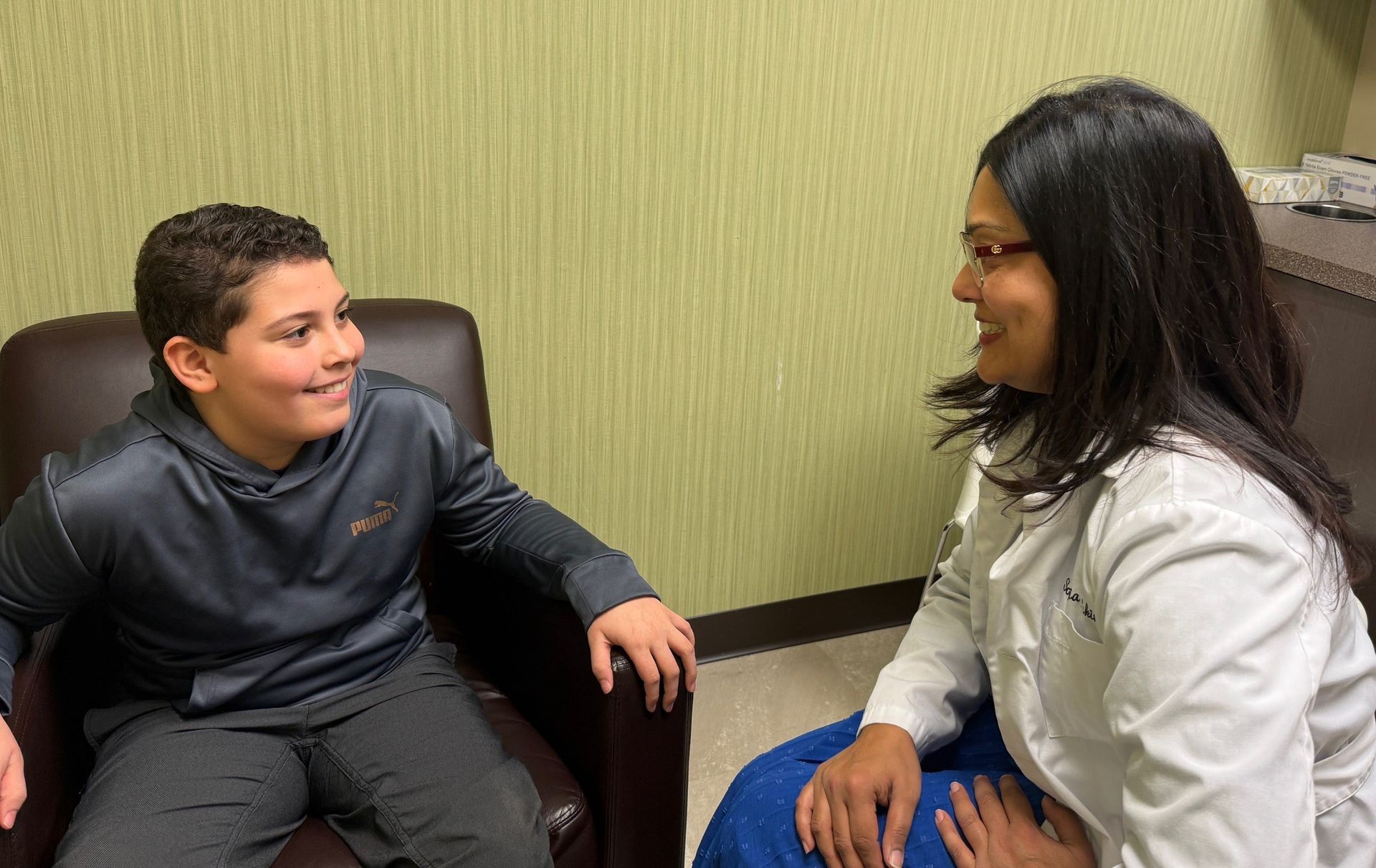 A person in a white coat smiles at a boy in a gray hoodie seated in a brown chair; both are indoors.