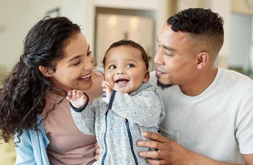 Smiling baby in grey onesie with parents, indoors.